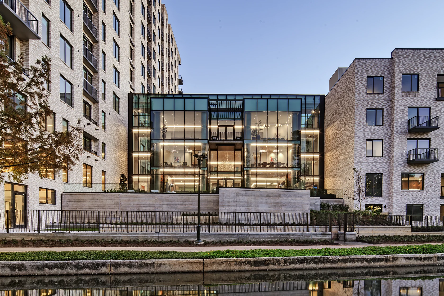 Modern residential complex featuring a glass-walled amenity building framed by brick apartments and reflected in the water.