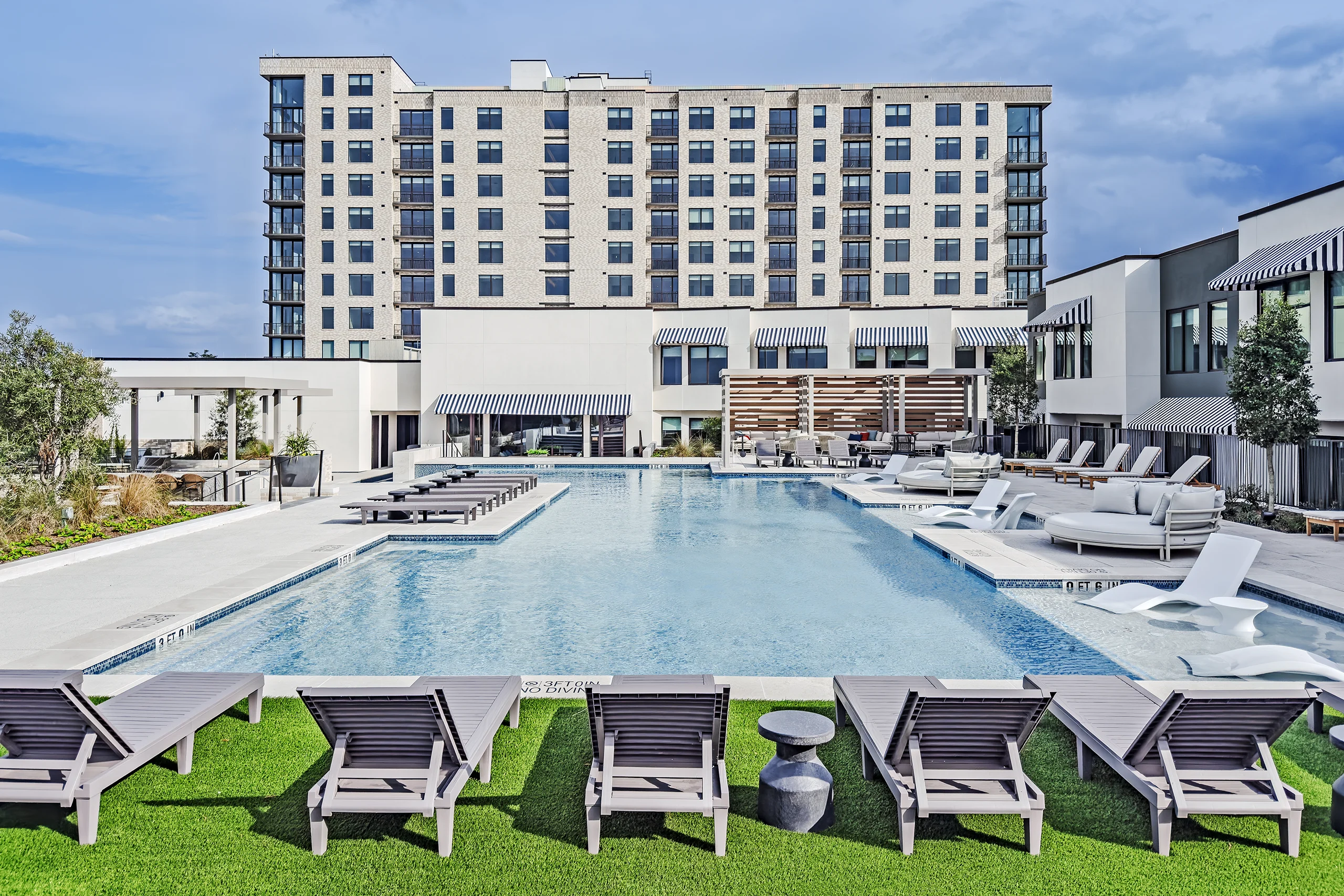 Resort-style outdoor swimming pool with rows of lounge chairs, landscaped grounds, and a modern multi-story residential building.