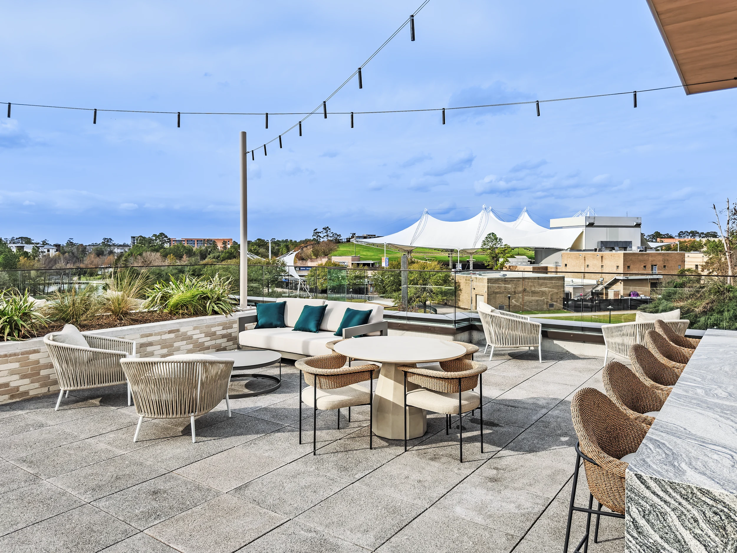 Rooftop terrace with cushioned lounge seating, woven chairs, dining tables, bar seating, string lights, and views of the surrounding landscape.